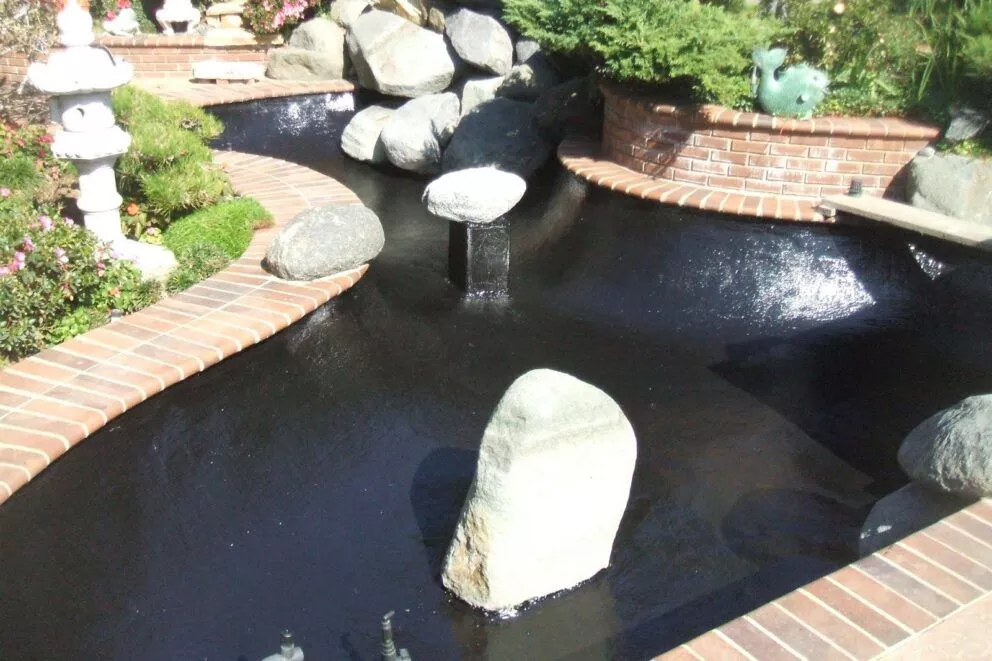 Black polyurea pond liner filled with water in a brick-edged koi pond with boulder accents