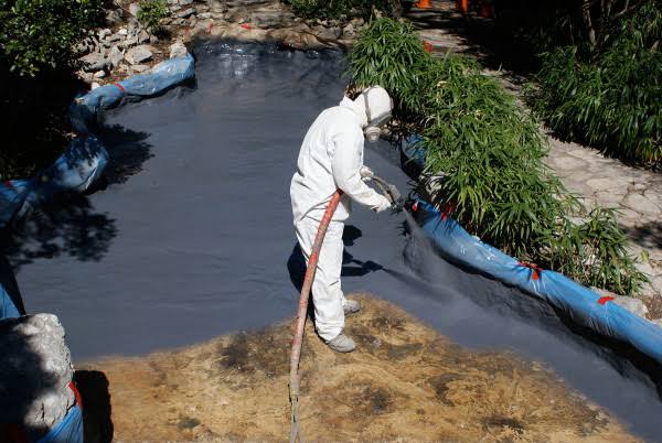 Contractor spray-applying polyurea aquatic coatings in a landscaped backyard pond with plants masked off
