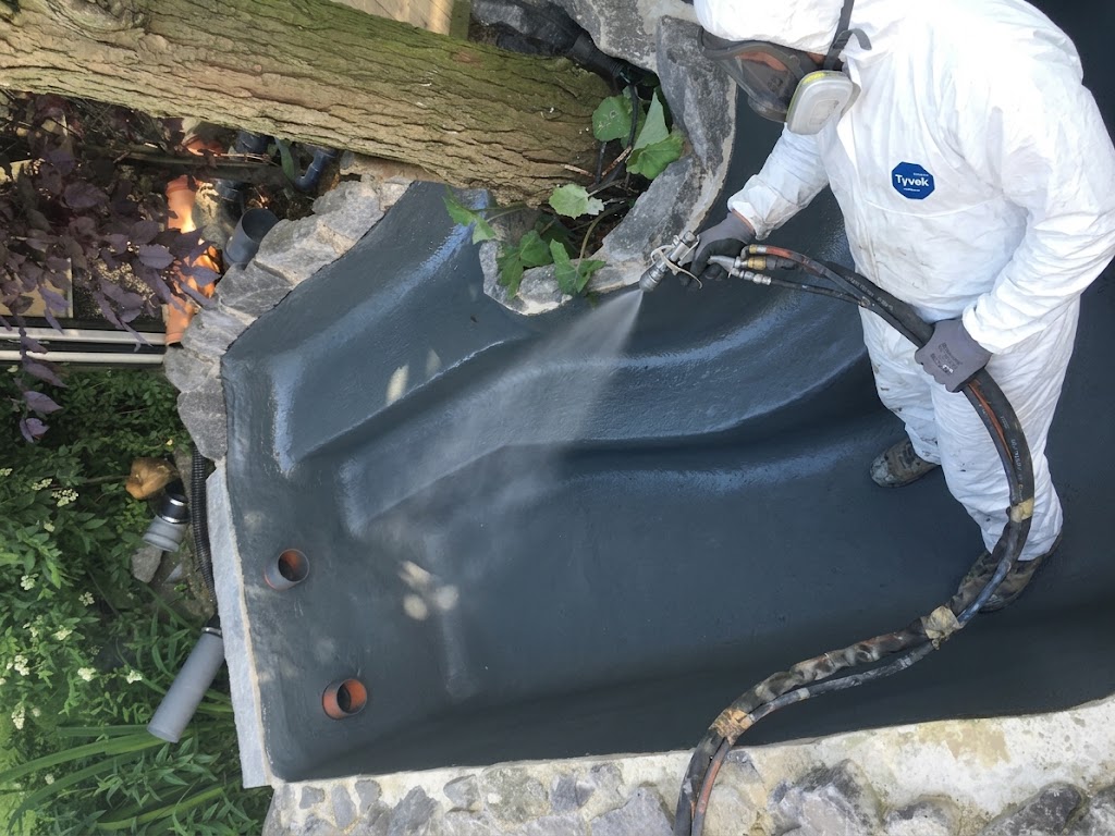 Applicator in Tyvek coveralls spraying polyurea aquatic coating over a stone stream and waterfall feature